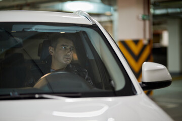 Man at steering wheel in modern car at underground parking, view through windshield.