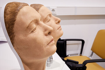 Two plastic masks imitating aged woman face, attached to stand on table in room, profile view.