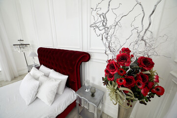 Bunch of artificial flowers, branches, berries in vase in bedroom with red bed.