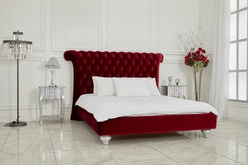 Interior of spacious white bedroom with large red bed, lamps, flowers, latticed window.
