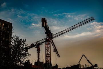 Tower crane on construction site at sunset. Silhouette against blue sky. Backlight forming silhouettes of trees and buildings. Construction activity in the foreground.
