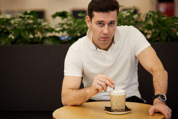 Man in white shirt sits at round table with cup of coffee-milk drink in club.