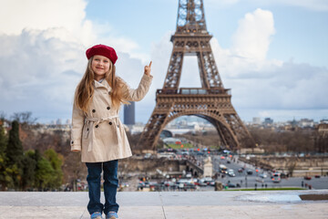 A happy, blonde girl in a trench coat and with a red beret hat stands in front of the Eiffel Tower in Paris, France