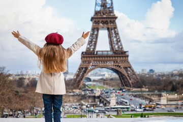 A happy tourist girl in a trench coat and with a red beret hat looks at the famous Eiffel Tower in Paris, France