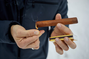 Close-up hands of man in jacket lighting tip of cigar with burning match.