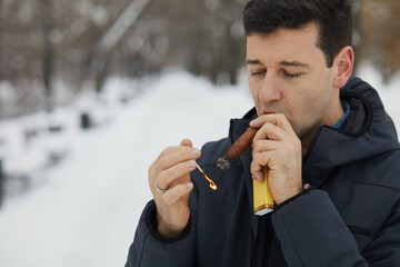 Man lights and puffs at cigar in winter snowy park.