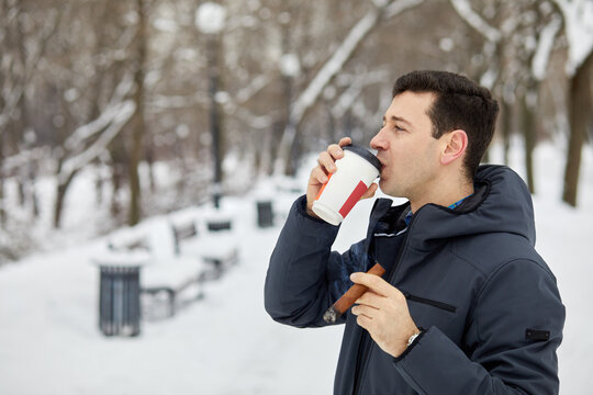 Man in jacket drinks coffee from paper cup and smokes cigar in winter park.