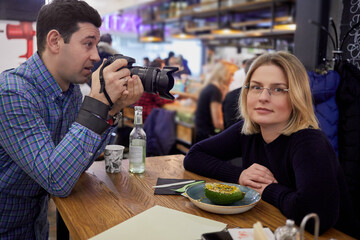 Man takes picture of woman sitting at table in cafe.