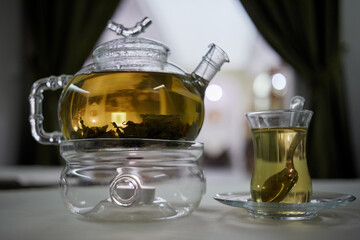 Transparent glass teapot on candle heater and glass teacup with teaspoon on table in cafe.