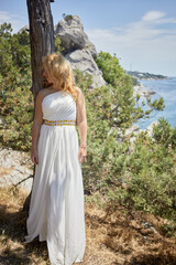 Blond woman in white dress with golden waist belt stands leaning her back to tree trunk with cliff and seashore at background on sunny day.