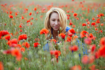 Blond woman in blue dress sits squatted on red poppies field.