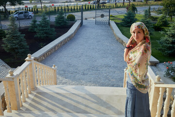 Woman in tatar head shawl stands on stairs outdoor.