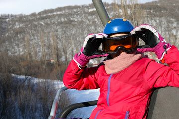 Happy girl in ski goggles looks away during lifting at cableway in ski resort at winter day