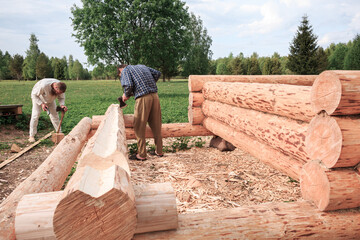 Two men are building a wooden house from logs in nature, cutting logs with axes, shavings, sawdust, logs in the foreground, construction, village, summer, greenery, forest, summer.