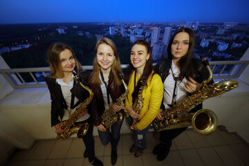 Four young women pose with wind instruments on rooftop in night city