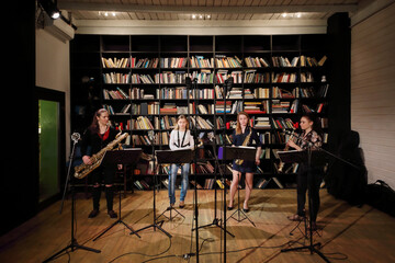 Four young pretty women play saxophone in room with bookshelves