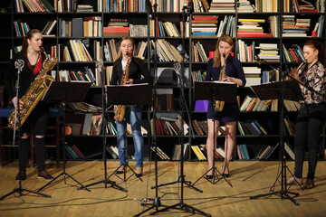 Four young pretty women play wind instruments in room with bookshelves