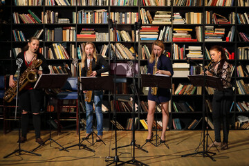 Four young pretty women with wind instruments perform in room with bookshelves