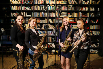 Four young pretty women pose with wind instruments in room with bookshelves