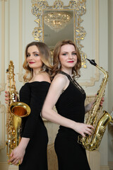 Two women in black stands with saxophones in baroque studio with mirror - obrazy, fototapety, plakaty