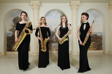 Four women in black dresses stand with saxophones in studio with pilasters - obrazy, fototapety, plakaty