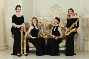 Four women in long dresses smiles with saxophones near couch in studio