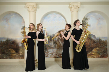 Four women in black dresses play saxophones in studio with pilasters - obrazy, fototapety, plakaty