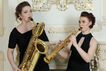 Two women in long dresses play saxophones in baroque studio with molding