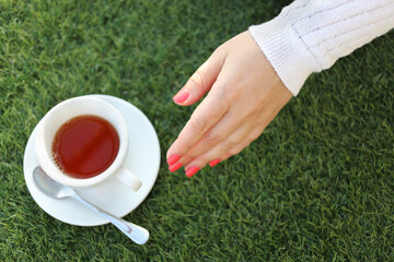 Female hand and white cup of hot tea on green lawn outdoor, top view