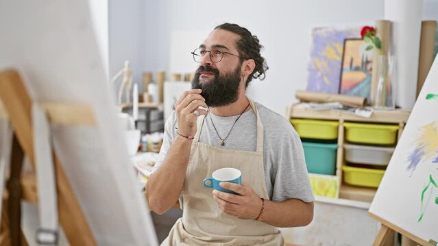 A thoughtful man with a beard holds a cup in an art studio, reflecting on his painting while wearing glasses and an apron.