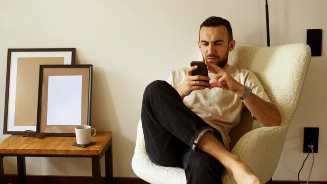 bearded guy sitting in cozy chair near floor lamp in cinema on white wall background near picture frames holding phone laughing and enjoying and wondering.