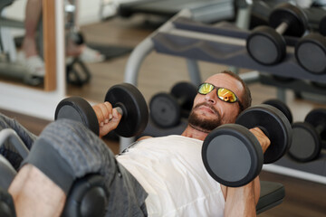 A bearded caucasian man in a gym lying on a bench and lifting dumbbells. Sportsmen wearing a white shirt, sunglasses and dark shorts. Various pieces of gym equipment are visible in the blur background