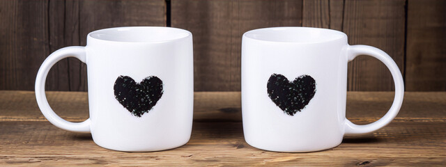 Two white coffee mugs with black heart-shaped prints placed on a wooden table against a wooden background.