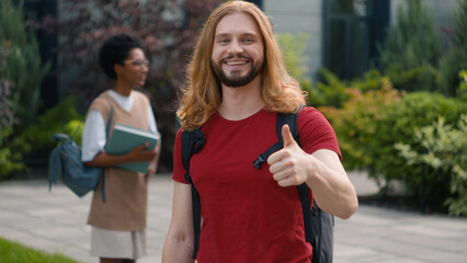 Caucasian young student man guy male showing thumb up agree recommend gesture approve like sign at camera standing against talking diverse multiracial students friends education conversation outdoors