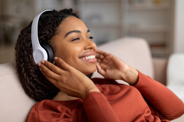 Relaxed young black woman wearing wireless headphones lying back on sofa