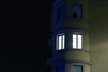 Fototapeta na wymiar A lone unrecognizable figure looks out from a brightly lit window at night smoking a cigarete
