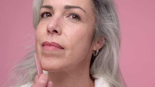 A woman with gray hair touches her cheek with gua sha while smiling in the mirror