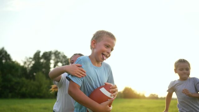 Happy team of children in the park in summer. Children run with a rugby ball. Children play outdoors on green grass. A team of children runs with a rugby ball in nature in the park. Team games concept