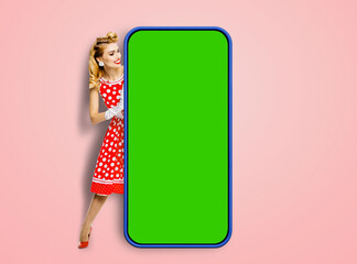 Full body woman in pinup dress hold show big size smartphone cellphone with mock up green chroma key screen. Pin up girl with mobile cell cellular phone, isolated against pink background.  Ad concept.