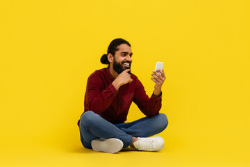Cool bearded indian guy sitting on floor, using smartphone
