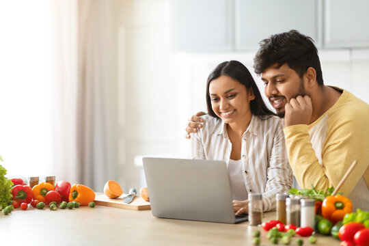 Happy Loving Young Indian Spouses Using Laptop While Cooking