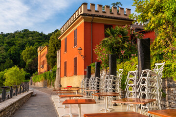 Cafe tables outside on embankment with colorful villa on Como lake in outdoor restaurant with nobody, Lenno comune, Lombardy, Italy. Popular travel and tourist destination on summer vacations - 762371745