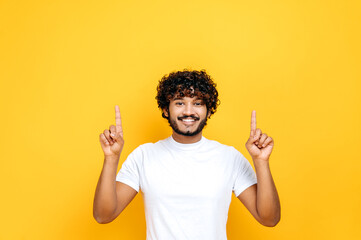 Happy positive indian or arabian curly-haired guy in basic white t-shirt, pointing fingers up at...