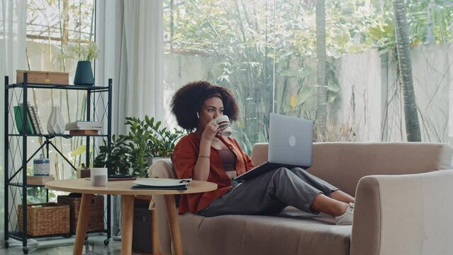Wide shot of young businesswoman analyzing sales using laptop while sitting on sofa at home and drinking coffee