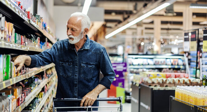 Mature Caucasian man is shopping in a supermarket, leaning against his cart with a smile on his face and taking a look at one of the shelves.