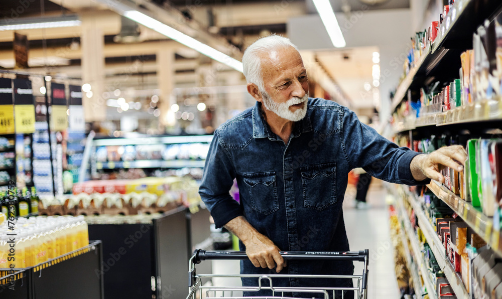 Wall mural retired man buying groceries. mature male buyer shopping groceries in supermarket from shelf standin