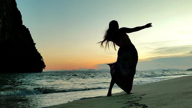 Young woman dancing against the light on the beach. Woman dancing while the sun goes down in the sea. Bellydancer. Silhouette of woman dancing on the beach while the sun goes down
