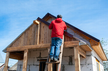 Fototapeta na wymiar A worker builds a roof in a house while standing on a wooden ladder. Blue sky