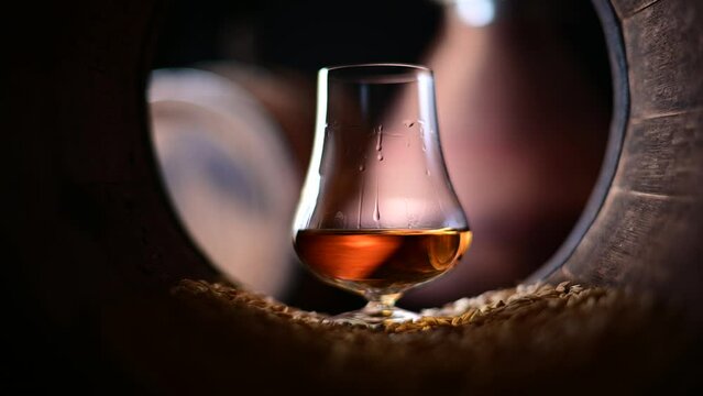 Drops flow down a glass of bourbon whiskey standing in an old oak barrel filled with barley grain. Copper alambic on background. Traditional alcohol distillery concept