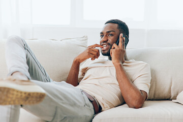 Happy African American man sitting on a black sofa at home, confidently holding his smartphone and...